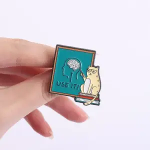 Emaille-Anstecker mit Katze, Brille und Gehirn-Motiv auf türkisem Schild, zwischen den Fingern gehalten